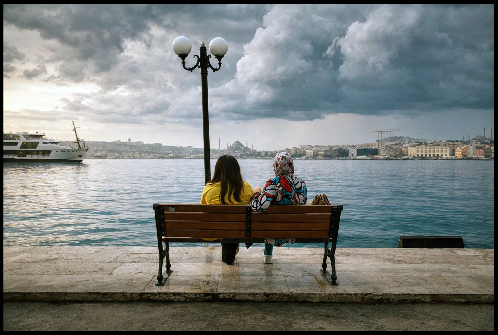 Two women seated on a bench facing the water beneath a cloudy sky