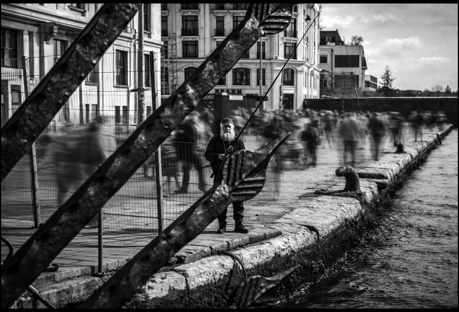 A monochrome waterfront scene with an angler and blurred figures walking past