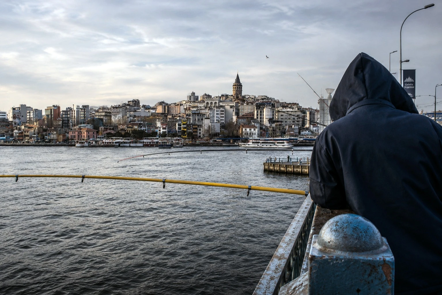 A hooded person fishing with the city across the water