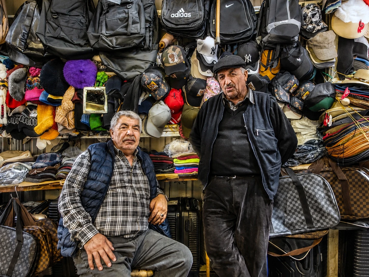 Two men at a market stall surrounded by hats and bags
