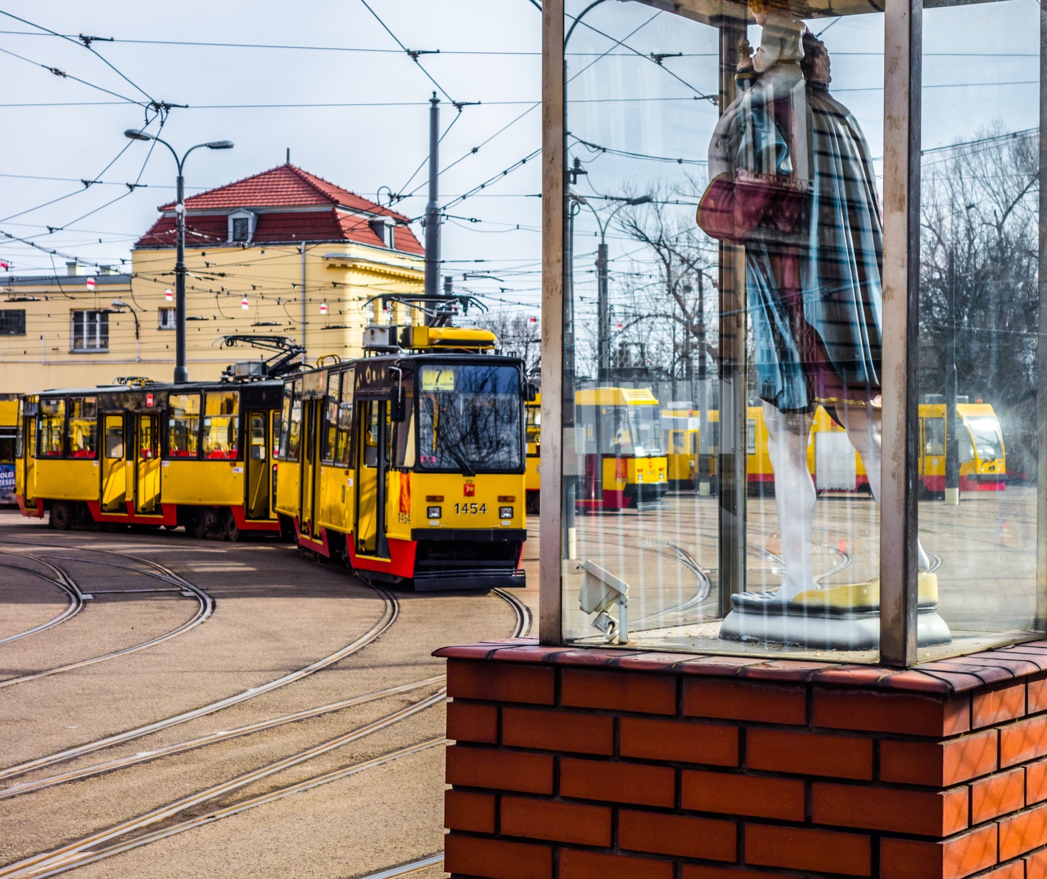 A roadside shrine with yellow and red trams passing behind it